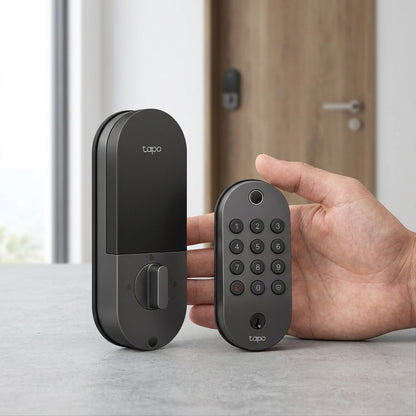 Scale comparison image shows the specific dark gray TP-Link Tapo Smart Wi-Fi Door Lock, with its detailed keypad and interior turnpiece, placed alongside a standard, realistic adult hand for an instant visual size reference within a clean room setting.
